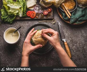 Female women hands holding homemade tasty burger on rustic kitchen table background with ingredients, top view