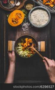 Female women hands holding pot and spoon and cooking pumpkin risotto on dark rustic kitchen table with ingredients, top view. Clean seasonal food and eating