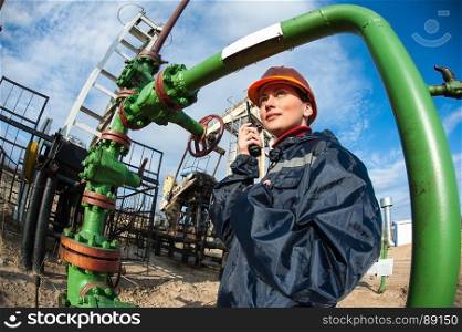 Female worker in the oil field talking on the radio wearing orange helmet and blue work clothes. Industrial site background.. Oil and gas industry worker.
