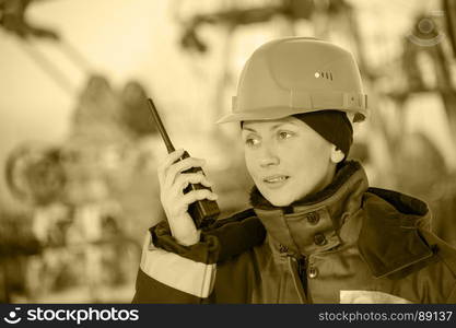 Female worker in the oil field talking on the radio wearing red helmet and blue work clothes. Industrial site background.Toned sepia.