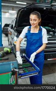 Female worker stands at the toolbox, car service. Vehicle repairing garage, woman in uniform, automobile station interior on background. Female worker stands at the toolbox, car service