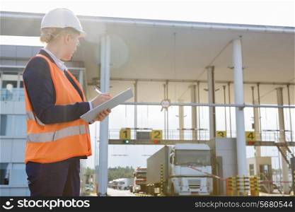 Female worker writing on clipboard while looking at trucks entering in shipping yard