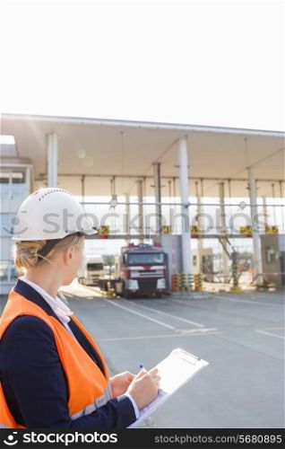 Female worker writing on clipboard while looking at trucks entering in shipping yard