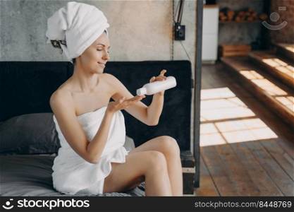 Feminine caucasian woman wrapped in towel is sitting on her bed. Girl applying body moisturizing cream squeezing it out from bottle after bathing. Morning shower and daily skin care at home.. Feminine caucasian woman applying body moisturizing cream. Morning shower and daily skin care.