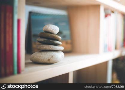 Feng Shui: Stone cairn at home in a book shelf, blurry books in foreground and background. Balance and relaxation. Sunlight.