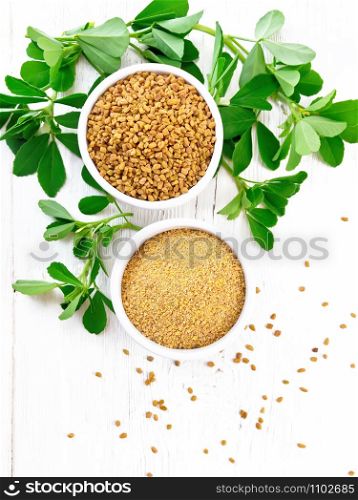 Fenugreek seeds and ground spice in two bowls and on a table with green leaves on wooden board background from above