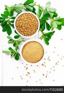 Fenugreek seeds and ground spice in two bowls and on a table with green leaves on wooden board background from above