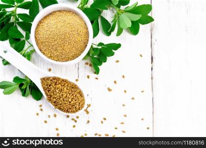 Fenugreek seeds in a spoon and ground spice in a bowl with green leaves on wooden board background from above