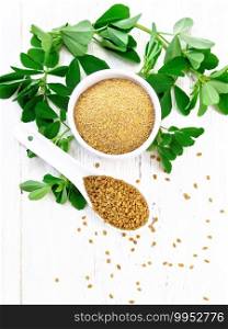 Fenugreek seeds in a spoon and ground spice in a bowl with green leaves on light wooden board background from above
