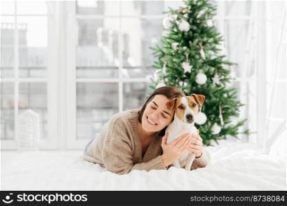 Festive mood, friendship, love between people and animals. Pleased smiling brunette woman in warm sweater hugs pet with love, enjoys spending time at home, prepares for Christmas celebration