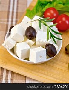 Feta cheese, olives, rosemary in a white bowl, tomatoes, green salad on the background of wooden boards and brown tablecloth