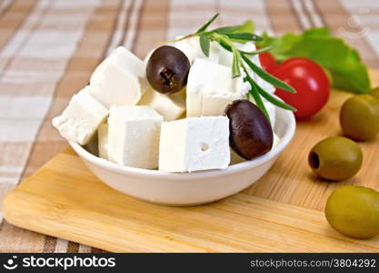 Feta cheese, olives, rosemary in a white bowl, tomatoes, lettuce on a wooden boards background and brown fabric