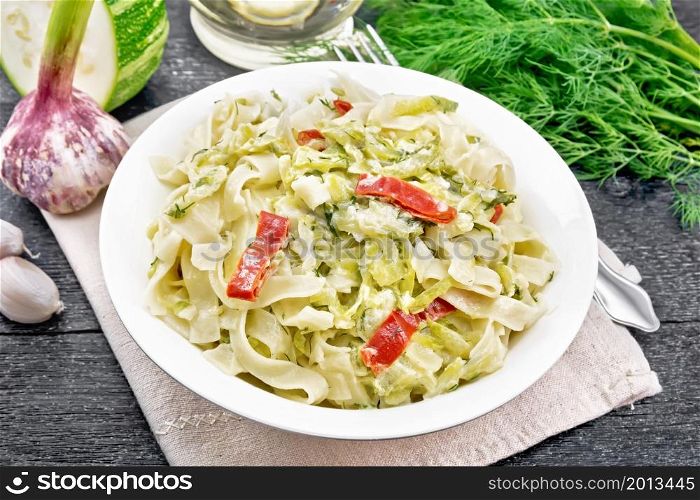 Fettuccine pasta with zucchini and hot red pepper in creamy sauce in white plate on a napkin, garlic and a fork on dark wooden board background