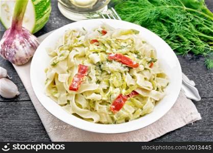 Fettuccine pasta with zucchini and hot red pepper in creamy sauce in white plate on a napkin, garlic and a fork on dark wooden board background