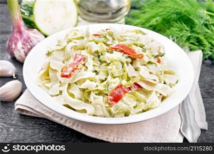 Fettuccine pasta with zucchini and hot red pepper in creamy sauce in white plate on a kitchen towel, garlic and a fork on black wooden board background