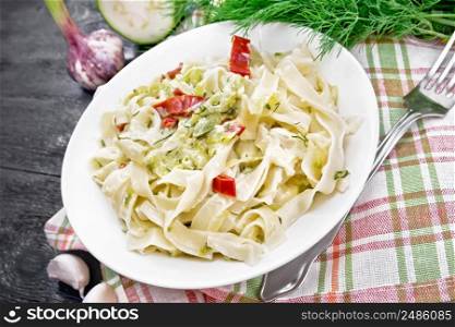 Fettuccine pasta with zucchini and hot red pepper in creamy sauce in white plate on a towel, garlic and a fork on wooden board background