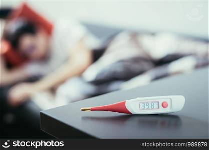 Fever thermometer in the foreground, ill man lying on the sofa in the blurry background