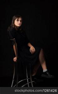 Fifteen-year-old girl sits on a bar chair, snasked on a black background in a dark