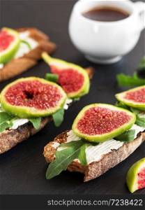 Figs on sliced toasted bread with cheese and arugula located on a black stone plate. A cup of strong espresso. Close-up.