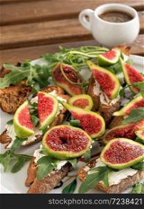 Figs on sliced toasted bread with cheese and arugula located on a white plate. A cup of strong espresso. Close-up. Wooden old background.