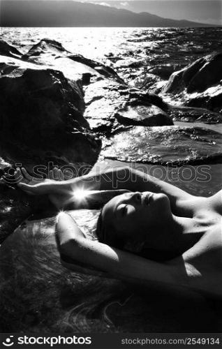Filipino young nude woman lying on back in water on rocky beach.