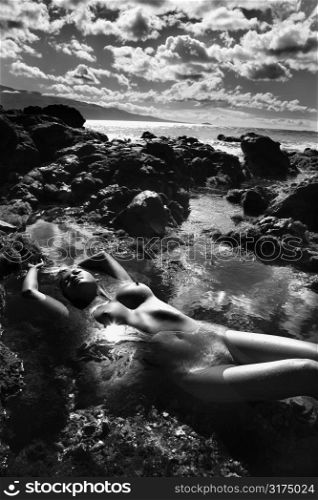 Filipino young nude woman lying on back in water on rocky beach with hands over head and cloudy sky.