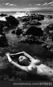 Filipino young nude woman lying on back in water on rocky beach with eyes closed and arms stretched over head.