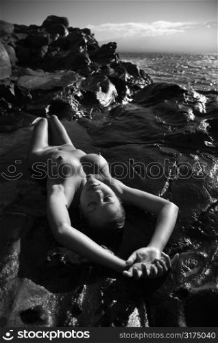 Filipino young nude woman lying on back in water with arms stretched over head on rocky beach.