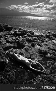 Filipino young nude woman lying on back with arms stretched over head on rocky beach.