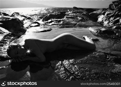 Filipino young nude woman lying on side in water on rocky beach.