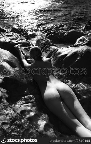 Filipino young nude woman lying on stomach in water on rocky beach.