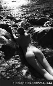 Filipino young nude woman lying on stomach in water on rocky beach.