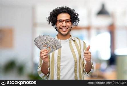 finance, currency and people concept - happy man holding hundreds of dollar money banknotes showing thumbs up over office background. happy man with money showing thumbs up at office
