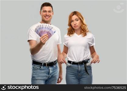 finance, saving and couple concept - happy young man in white t-shirt holding euro money and sad woman with empty pockets over grey background. couple with euro money and empty pockets