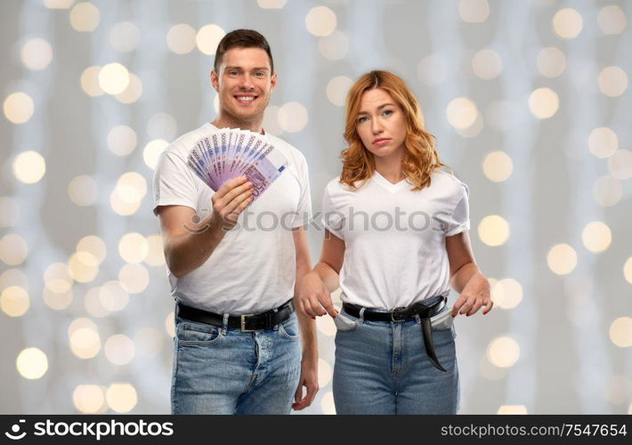 finance, saving and couple concept - happy young man in white t-shirt holding euro money and sad woman with empty pockets over festive lights background. couple with euro money and empty pockets