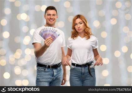 finance, saving and couple concept - happy young man in white t-shirt holding euro money and sad woman with empty pockets over festive lights background. couple with euro money and empty pockets