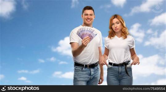 finance, saving and couple concept - happy young man in white t-shirt holding euro money and sad woman with empty pockets over blue sky and clouds background. couple with euro money and empty pockets