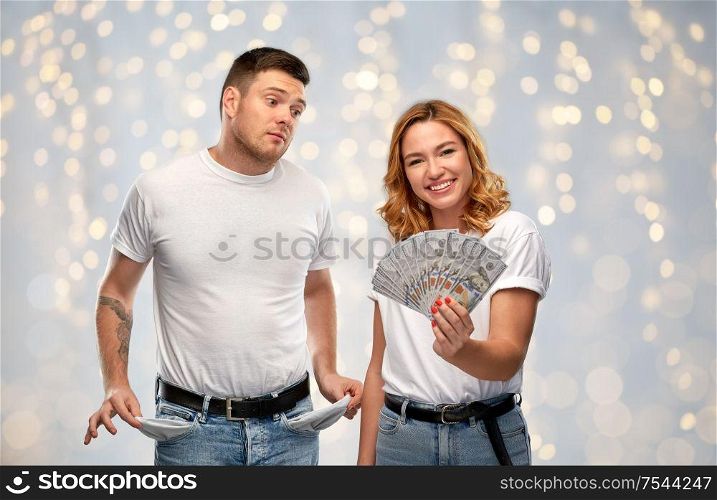 finance, saving and couple concept - happy young woman in white t-shirt holding dollar money and sad man with empty pockets over festive lights background. woman holding money and sad man with empty pockets