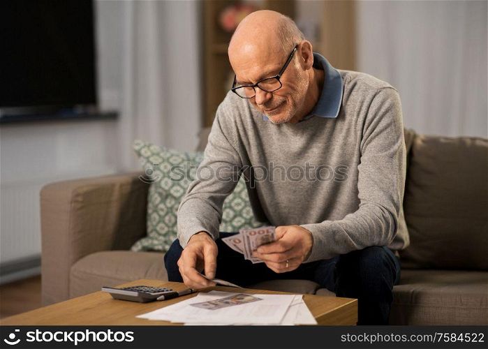 finances, savings and people concept - smiling senior man with calculator and bills counting money at home in evening. senior man counting money at home