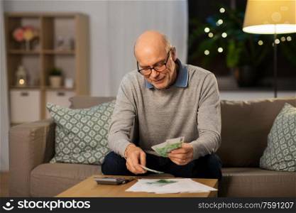 finances, savings and people concept - smiling senior man with calculator and bills counting money at home in evening. senior man counting money at home