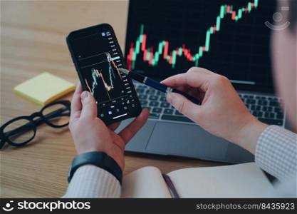 Financial chart on laptop screen. Man is pointing with pen to phone screen. Online trading with smartphone and computer through internet. PC on the desk at workplace in office. Business development.. Man is pointing with pen to financial chart phone screen. PC on the desk at workplace in office.