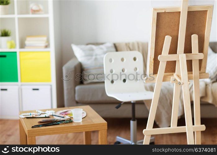 fine art, creativity and painting concept - wooden easel and artistic tools at home room or studio. easel and artistic tools at home or art studio