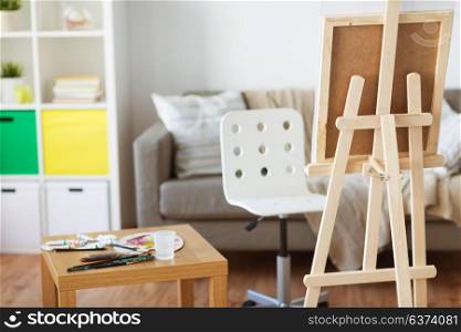 fine art, creativity and painting concept - wooden easel and artistic tools at home room or studio. easel and artistic tools at home or art studio