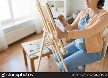 fine art, creativity and people concept - close up of artist with easel and paintbrush painting at studio. artist with easel and brush painting at art studio. artist with easel and brush painting at art studio