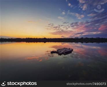 Fine art shot with an old, rusty catamaran frame forgotten on the pond. Scenery landscape, peaceful sunset sky background with colorful clouds reflection on the lake water.