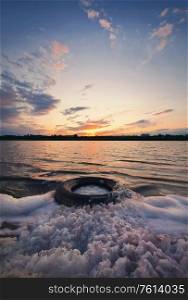 Fine art vertical shot, old vehicle tire thrown on the shore of a pond, covered with fluffy surf foam created by waves. Scenery landscape, peaceful sunset sky background over the city on the horizon.