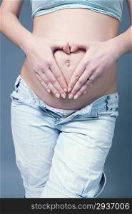 Fingers in heart shape over navel on pregnant white woman&acute;s belly. Wearing light blue top and jeans. Pregnancy collection.