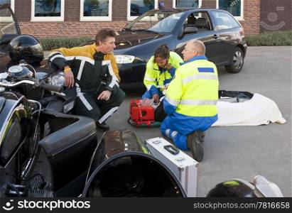 Fireman talking to a paramedic at the site of an accident, whilst a female paramedic checks up on the victim on a brancard