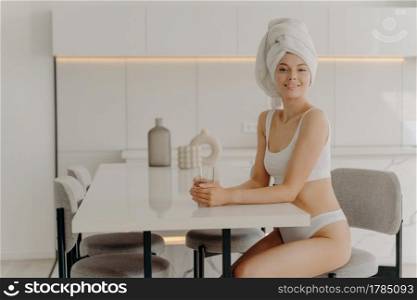 First thing in the morning. Relaxed slender woman sitting in stylish kitchen after taking shower with glass of pure mineral water, wears white underwear and towel over wet hair. Weightloss and dieting. Relaxed beautiful slender woman sitting in kitchen after taking shower with glass of water