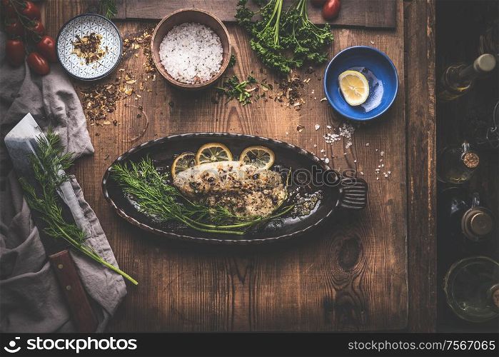 Fish fillet with lemon and herbs in backing pan on rustic kitchen table background with ingredients. Top view